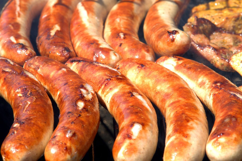 Close-up of ten sausages on a grill. The sausages have been browned, and some have begun to split. and another cut of meat is seen top right.
