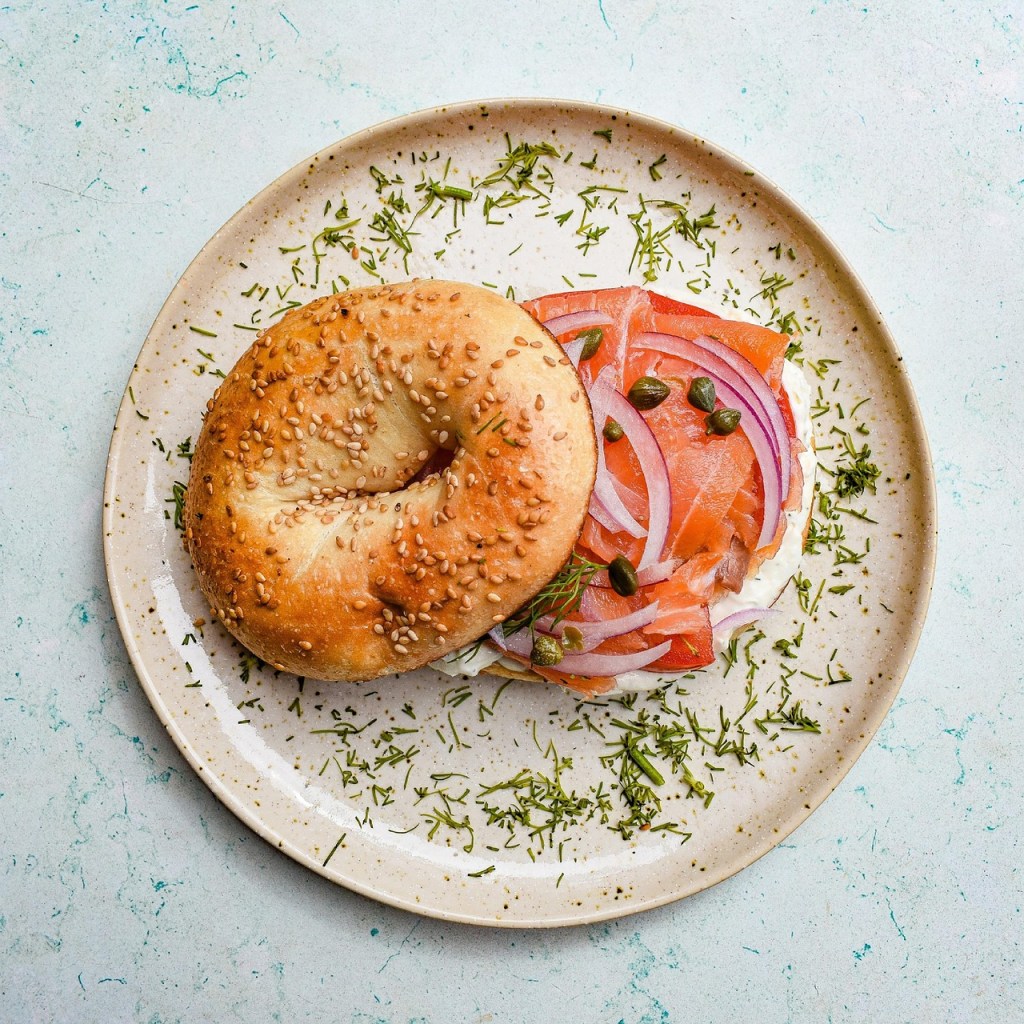 A sesame bagel topped with cream cheese, smoked salmon, red onion, and capers, garnished with herbs on a light-colored plate.
