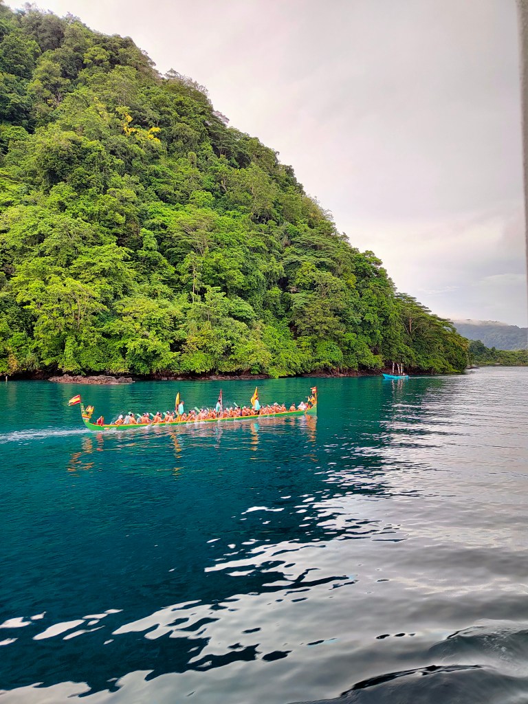 A traditional boat full of people on water with a tropical jungle backdrop. 