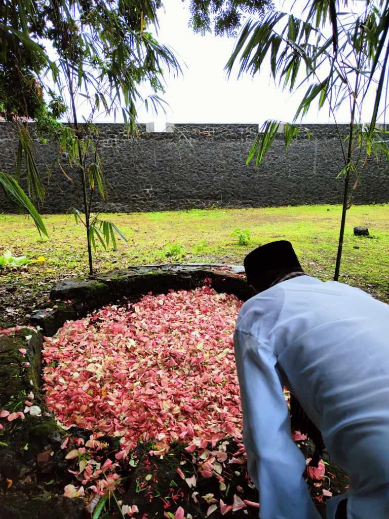A person in traditional clothing leans over a stone well surrounded by a grassy area, covered with pink flower petals, under a canopy of trees.