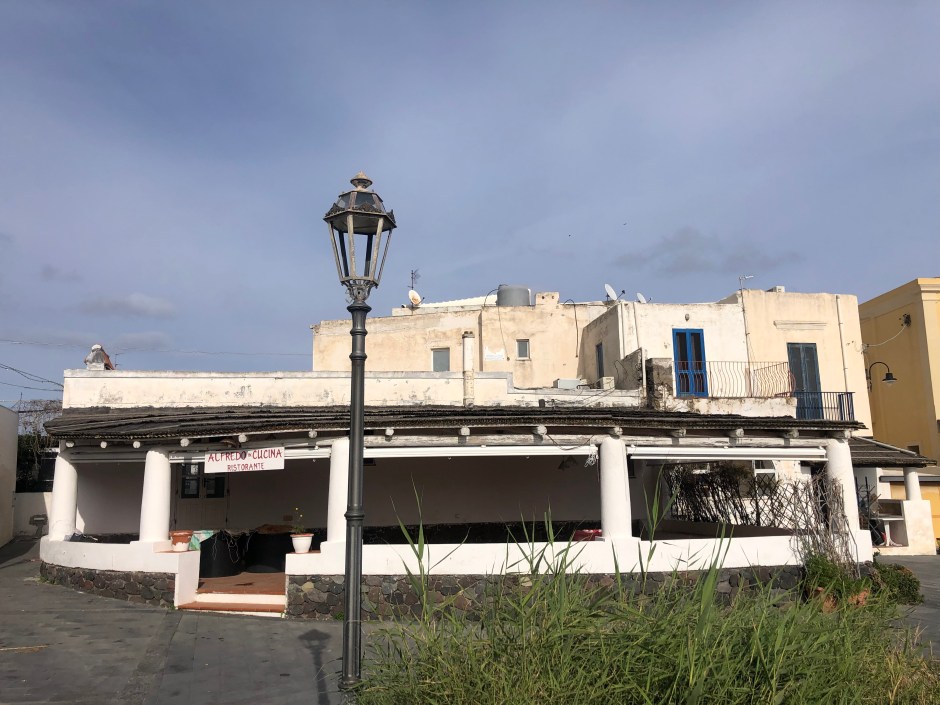 Building behind the dirt field, used for drying fish in case of bad weather. This is the structure that Elbano Tobia transformed into his pizzeria, La Marinara, and which today is the famous Ristorante da Alfredo, as stated on the hanging sign. Photo taken during the research residency Gastronomic and Cultural Adaptation to Climate Change in Mediterranean Island Areas: An Ecocritical and Participatory Analysis, at Fondazione Studio Rizoma, Palermo, Italy.