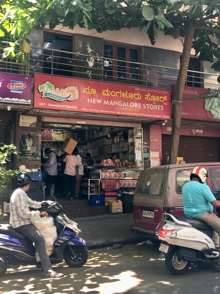 Exterior view of New Mangalore Stores, a local grocery shop in Bengaluru, featuring a vibrant sign and customers shopping for Mangalorean food items.