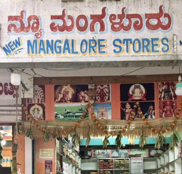 Entrance of a Mangalore Store, featuring a sign that reads 'New Mangalore Stores' in both Kannada and English, adorned with traditional decorations and images of deities.